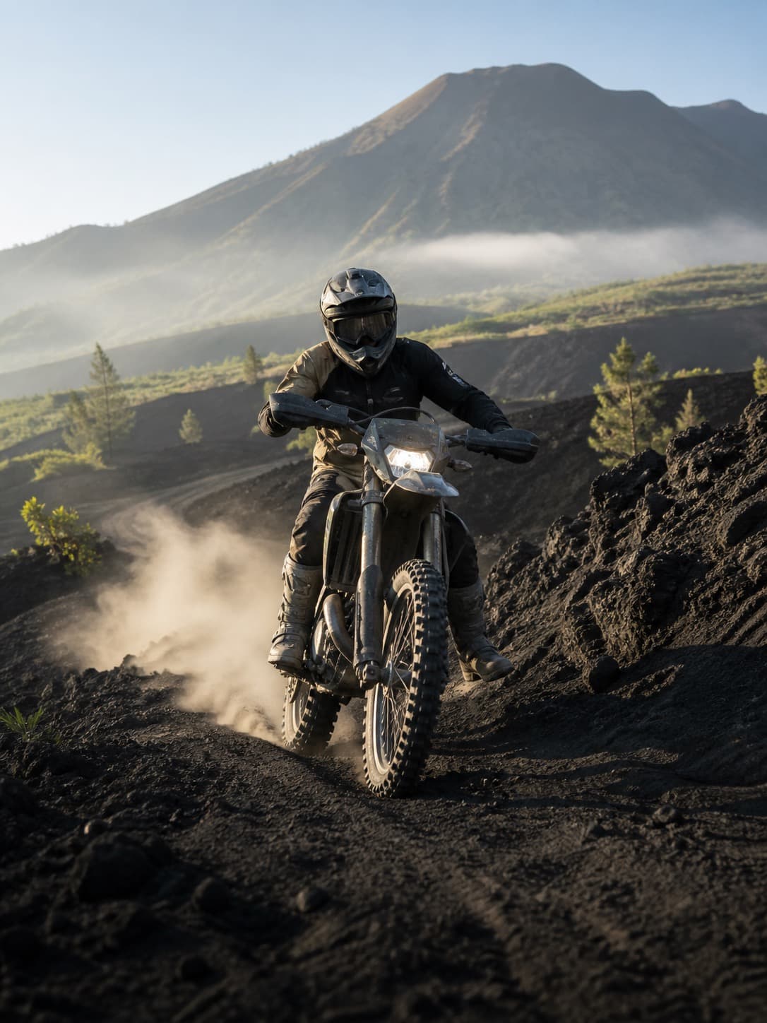 A helmeted rider on a trail bike throwing up a plume of ash, Mount Batur behind